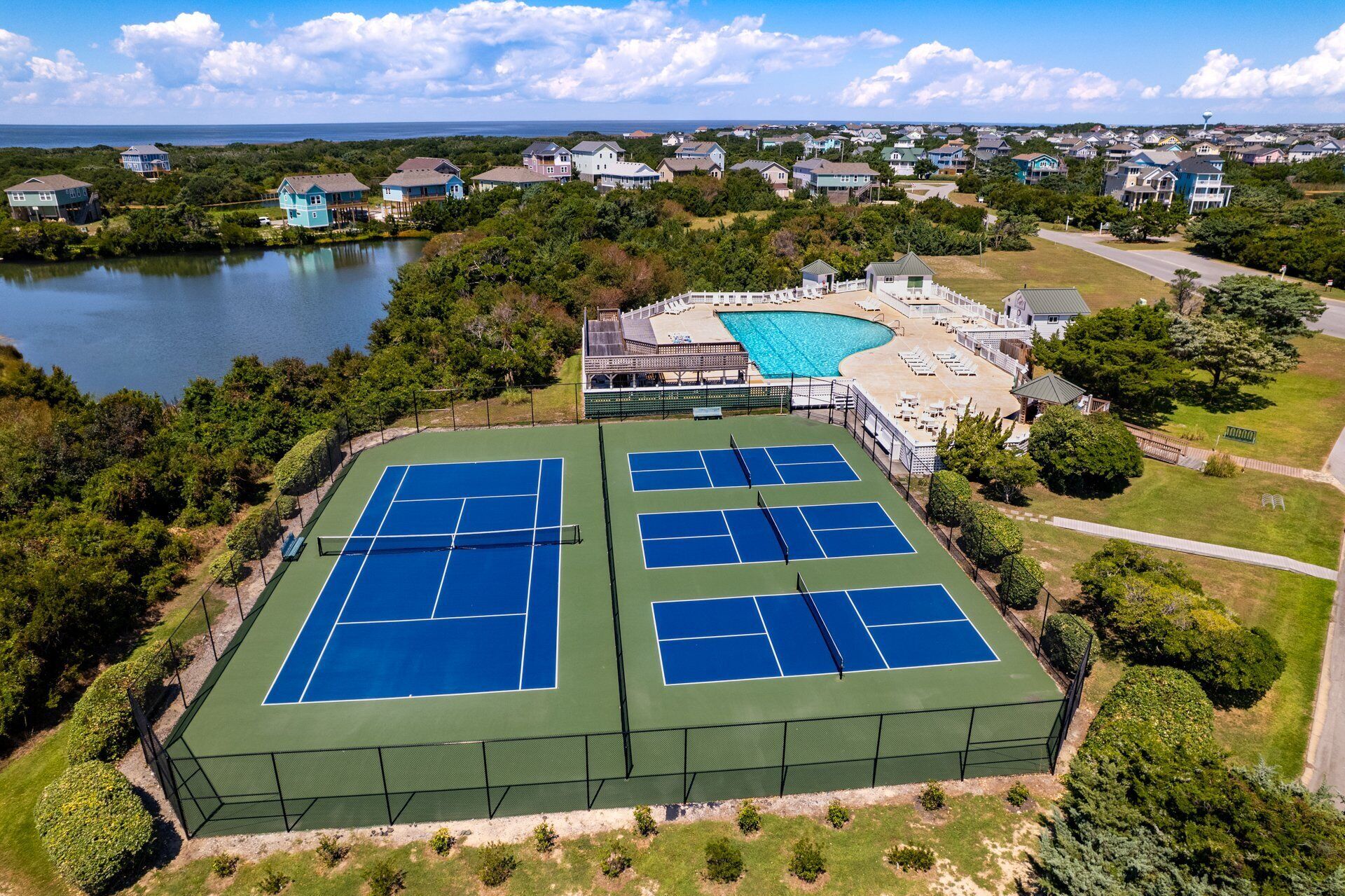 A tennis court complex with multiple courts, surrounded by residential houses and a lake.