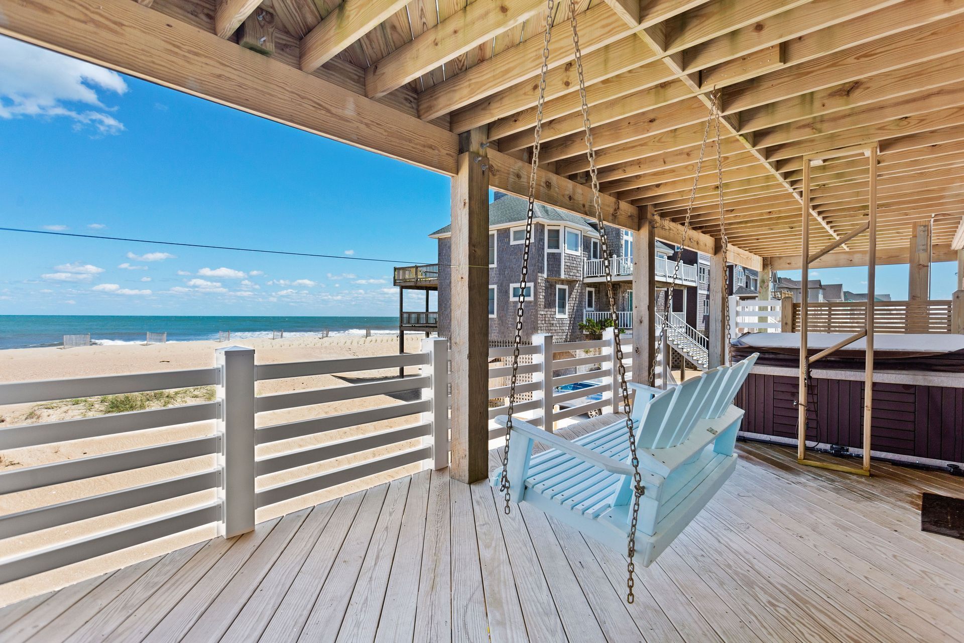 A wooden deck with a swing, overlooking a beach and a building with multiple balconies.