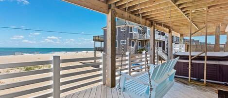 A wooden deck with a swing, overlooking a beach and a building with multiple balconies.