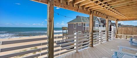 A wooden deck with a view of the beach and ocean, featuring a house with a balcony and a fence.