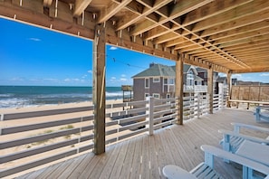 A wooden deck with a view of the beach and ocean, featuring a house with a balcony and a fence.