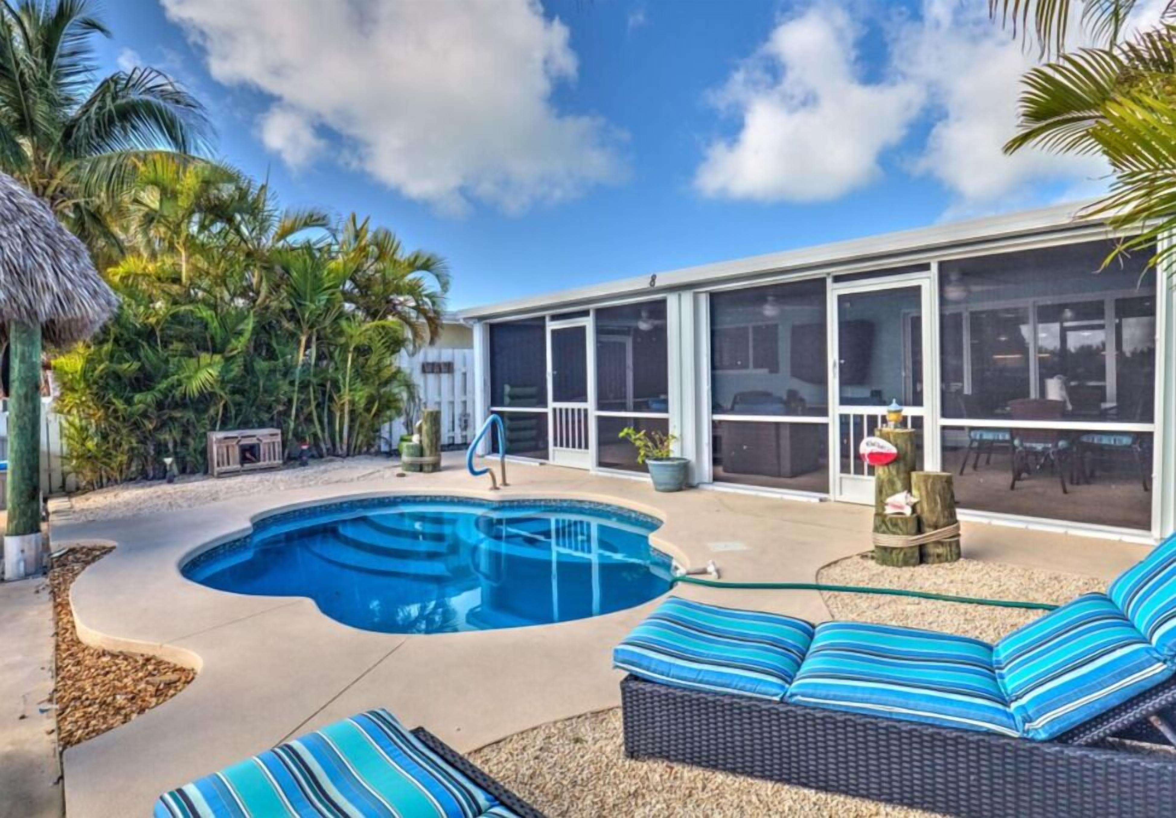 A modern outdoor pool area with a curved design, surrounded by tropical plants and a covered patio with seating.