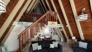 Een loft-achtige woonruimte met een houten trap, twee donkere banken, een rieten salontafel en een eethoek op de achtergrond.