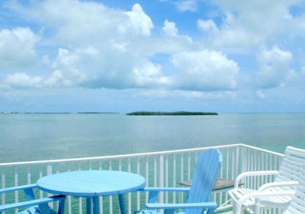 A deck with a round table and two chairs, overlooking a calm sea with a small island in the distance.