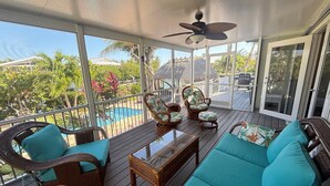 Un área de estar al aire libre con techo, con mobiliario de mimbre y una mesa de centro de vidrio, con vistas a una piscina y vegetación tropical.