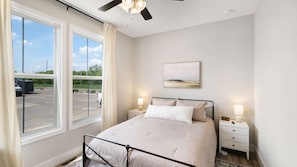 A bedroom with a bed, two bedside tables, a ceiling fan, a window with curtains, and a painting on the wall.
