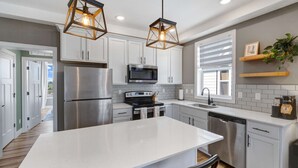 Une cuisine moderne avec des meubles blancs, des appareils en acier inoxydable, un îlot central, des luminaires suspendus et une fenêtre avec des stores.