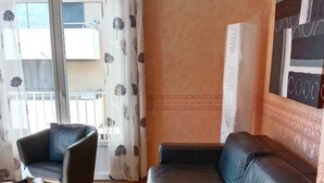 Ein Zimmer mit einer schwarzen Ledersofa, einem passenden Sessel und einem kleinen Tisch. Es gibt einen Balkon mit einem weißen Vorhang mit floralem Muster.