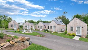 Une rangée de cabanes en bois, chacune dotée d’une porte numérotée, une allée pavée et un petit espace aménagé avec un foyer.