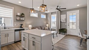 Cocina moderna con una isla central, comedor con mesa y sillas, ventilador de techo y ventanas amplias que ofrecen una vista.