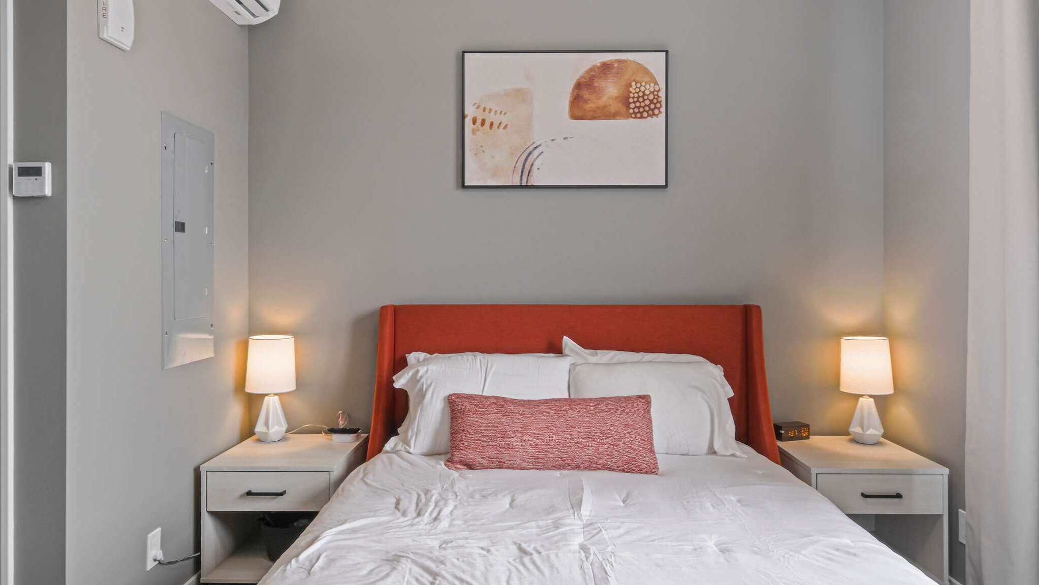 A hotel room with a bed, two bedside tables, a wall-mounted air conditioning unit, a framed artwork, and a digital clock.