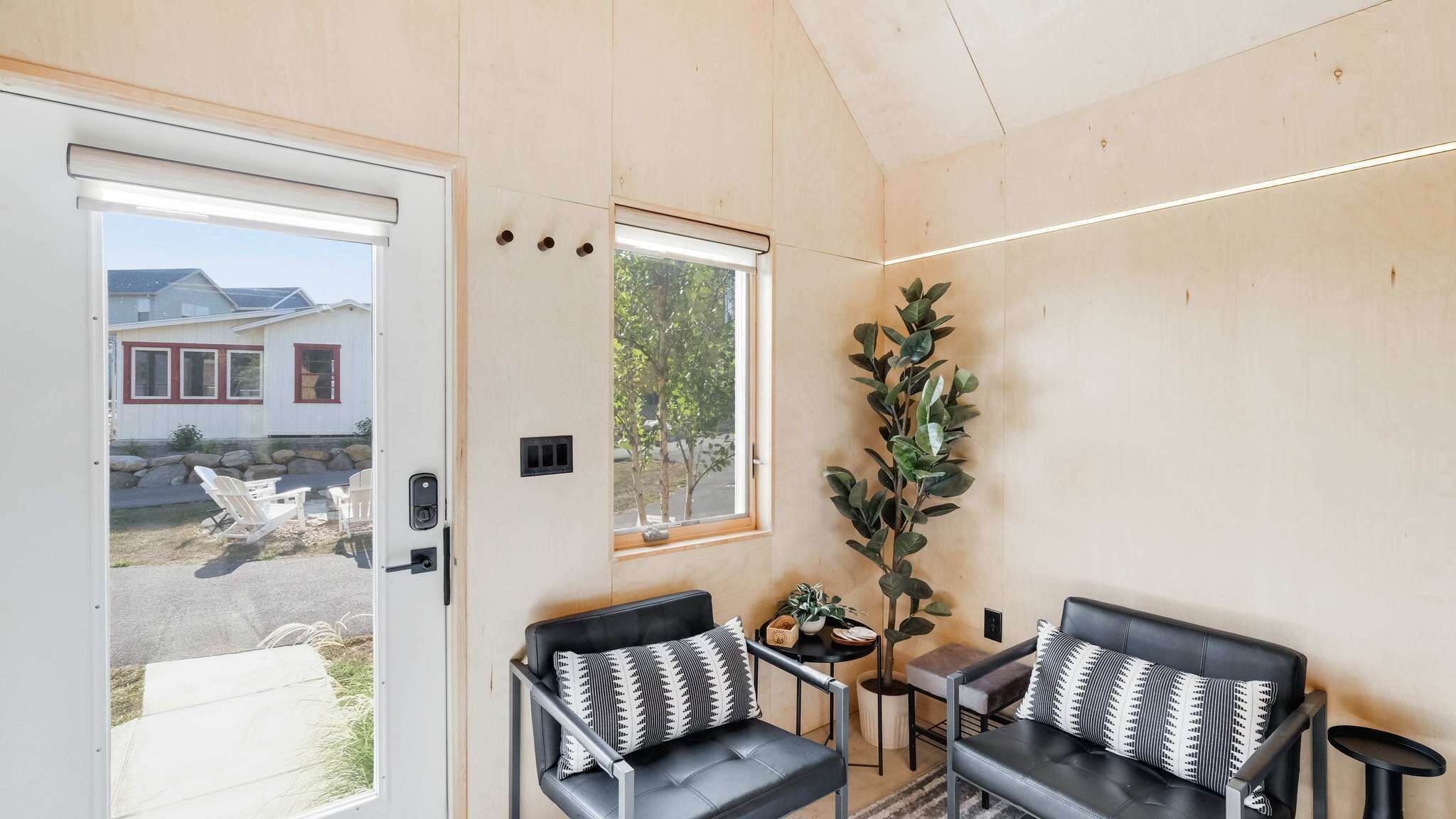 Une pièce de taille modeste dotée d’une porte coulissante, de deux canapés en cuir noir ornés de coussins à motifs, d’une petite table sur laquelle est posée une plante, et d’une fenêtre donnant sur une maison et des arbres.