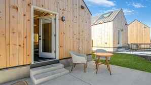 En træbygning med åben dør til et rum, et lille udendørs siddeområde med bord og stole, og en græsplæne.