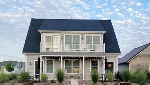 Una casa de dos pisos con balcón, un porche con asientos y un pequeño jardín.