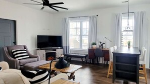 Ein Wohnzimmer mit Sofa, Sessel, Fernseher, Kamin, Esstisch und Deckenventilator.