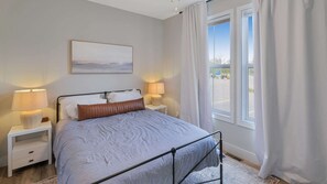 Een slaapkamer met een bed, twee nachtkastjes met lampen, een raam met gordijnen en een schilderij aan de muur.