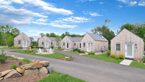 A row of wooden cabins with numbered doors, a paved driveway, and a small fire pit area. - The Bunkies @ Heritage Harbor (Ottawa)
