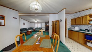 Uma área de refeições com mesa de madeira, cadeiras e um tapete verde. Há uma cozinha com armários de madeira e um micro-ondas.