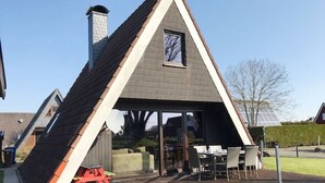 Een gebouw in een driehoekige vorm met een schuin dak, schoorsteen en een overdekt terras met tafels en stoelen.