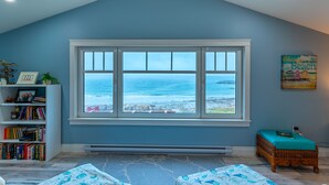 A room with a large window overlooking the ocean, a bookshelf, a wicker bench, and a framed picture on the wall.