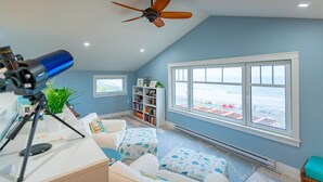 Una habitación con ventilador de techo, un telescopio, una estantería, una ventana con vistas a la playa y un rincón de descanso acogedor con cojines estampados.