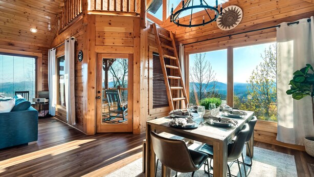 A wooden cabin with a dining area, a loft bedroom, and a view of the mountains.
