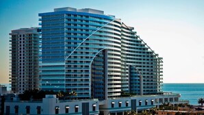 A modern high-rise hotel with multiple balconies, a curved facade, and a view of the ocean.