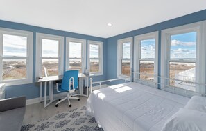 A bedroom with a large bed, a desk with a chair, and multiple windows offering a view of the beach and dunes.