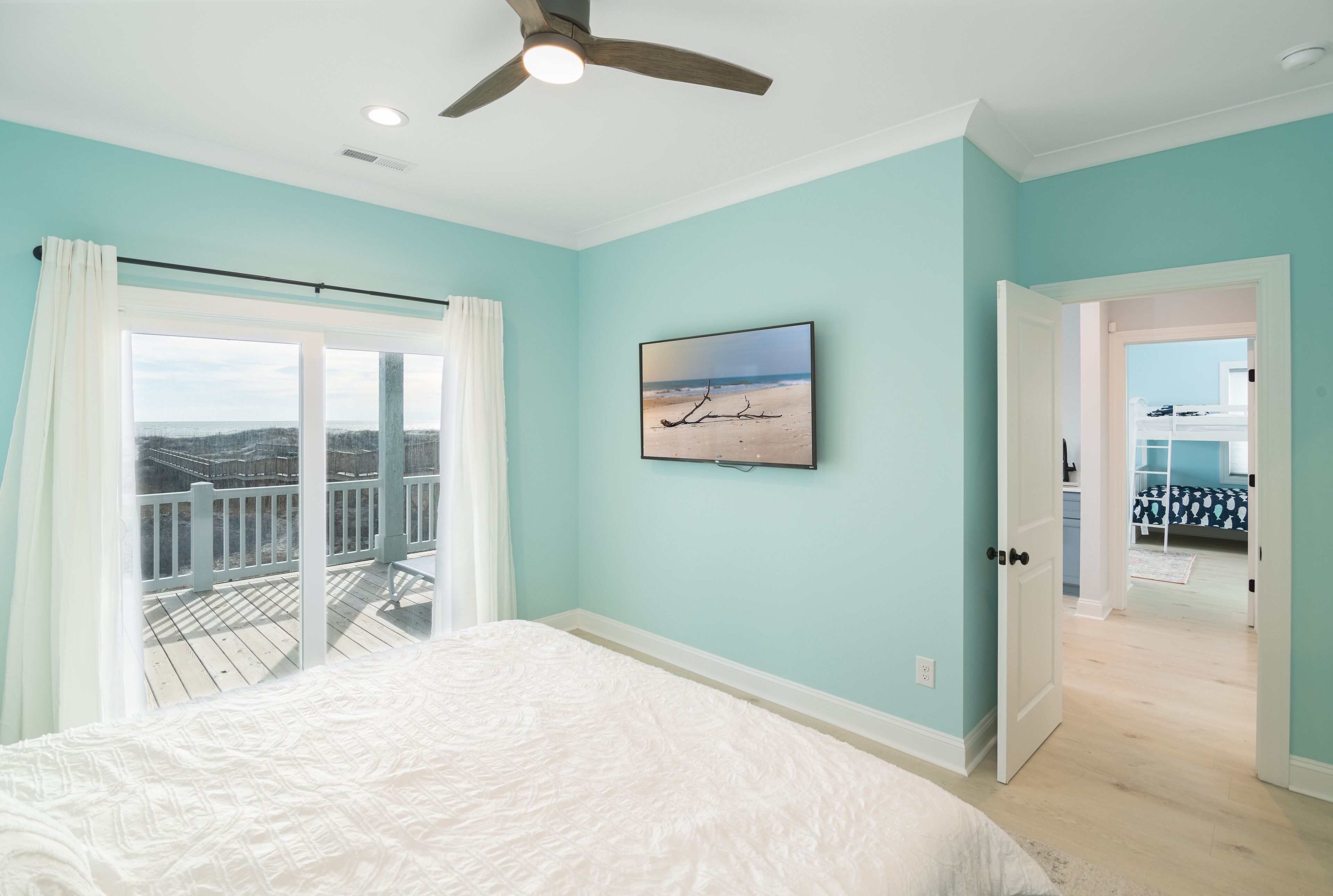 A bedroom with a large bed, a ceiling fan, a view of the balcony, and a door leading to another room.