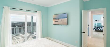 A bedroom with a large bed, a ceiling fan, a view of the balcony, and a door leading to another room.