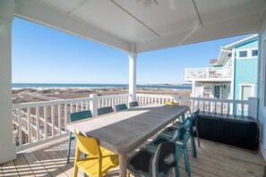 A covered outdoor area with a wooden table and chairs, a white railing, and a view of the beach and ocean.