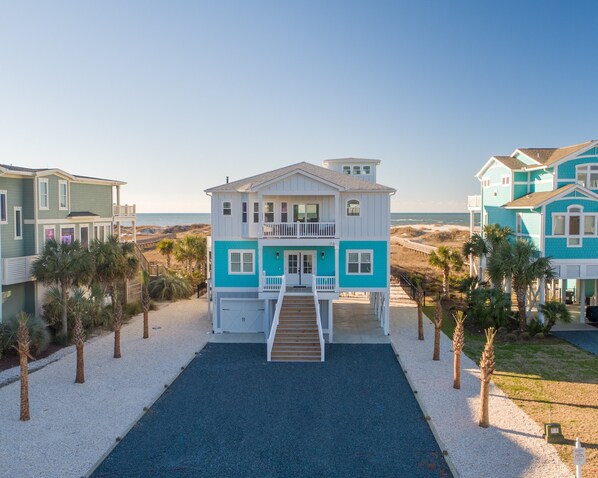 A two-story beachfront house with a balcony, a driveway, and a pathway leading to the entrance.