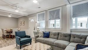 Ein Wohnzimmer mit einem blauen Sessel, einem grauen Sofa und einem weißen Hocker. Im Hintergrund ist ein Esstisch mit Stühlen zu sehen.
