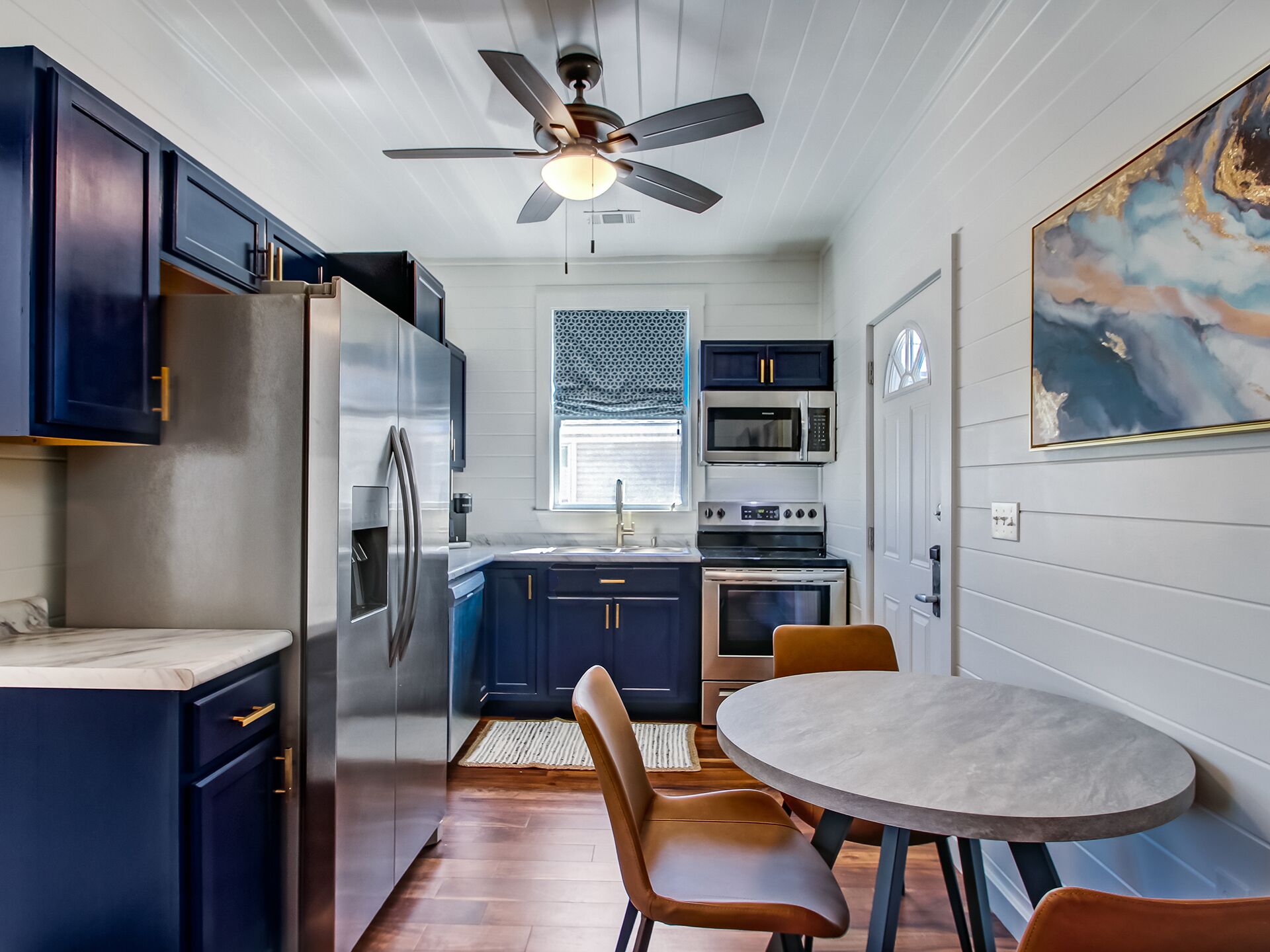 Cocina compacta con armarios azul oscuro, un refrigerador de acero inoxidable, un microondas y un horno. Hay una mesa de comedor con dos sillas.