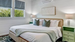 A bedroom with a bed, two bedside tables, a ceiling fan, an air conditioning unit, and a window with a view of greenery. - Multi-Unit Home, Steps to the Beach, Pool Access (Tybee Island)