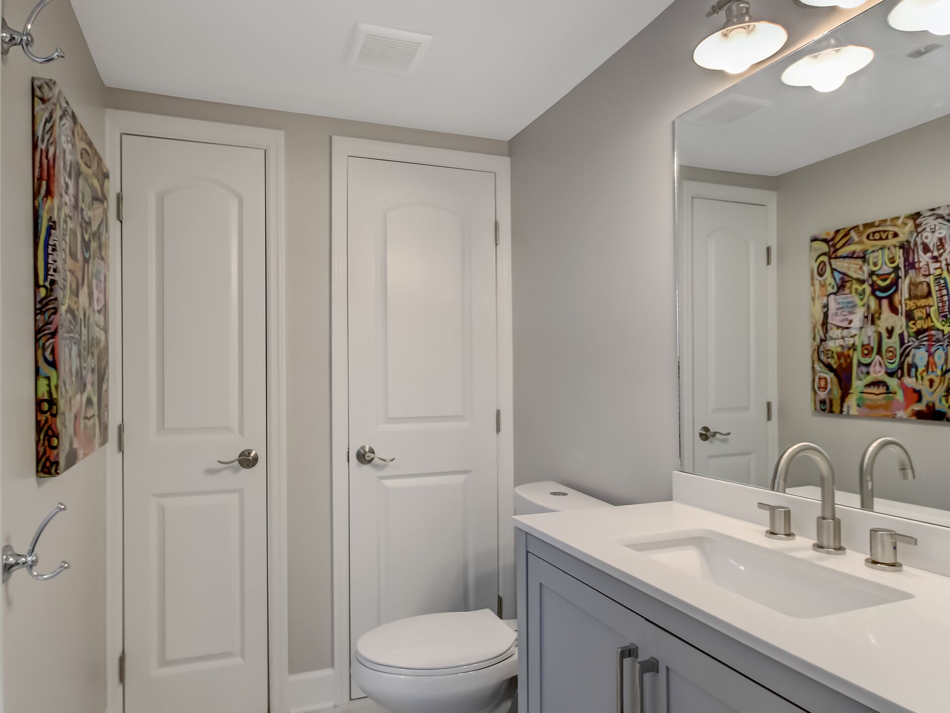 A bathroom with two white doors, a white toilet, a white sink with a mirror, and a colorful abstract painting on the wall.