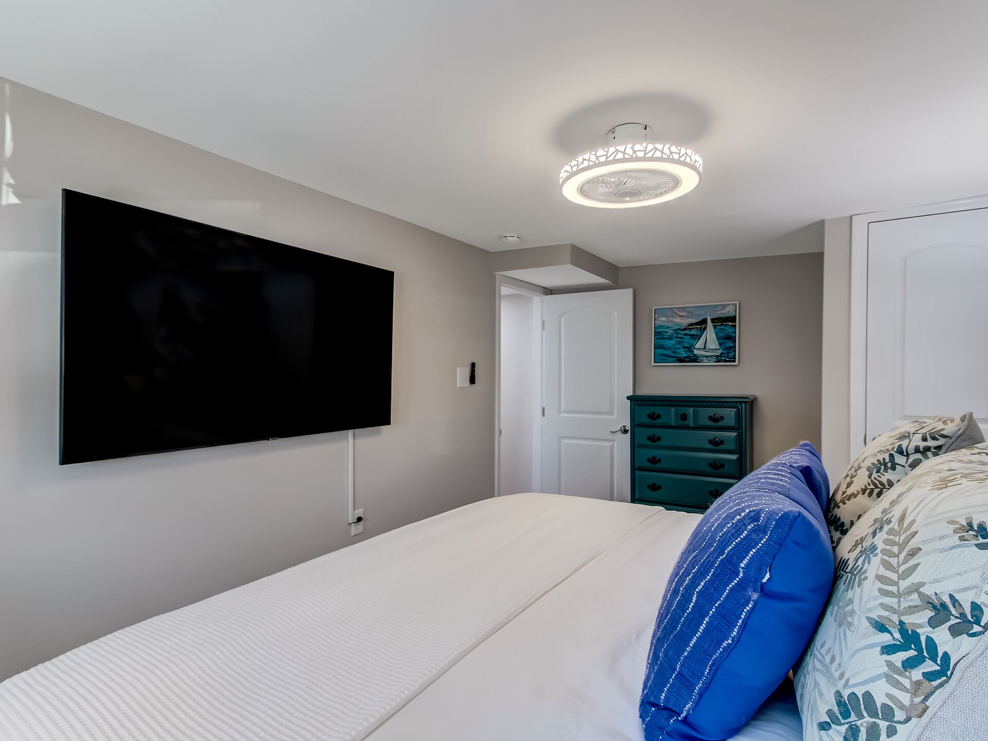 Een slaapkamer met een grote flatscreen televisie aan de muur, een bed met witte beddengoed en blauwe kussens, een blauwgroene dressoir en een plafondlamp.