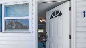 Una puerta blanca con un letrero con el número 1, una ventana con persianas y vista a una habitación con un cojín de cuadros y una silla azul.