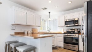 A modern kitchen with white cabinetry, a central island with stools, stainless steel appliances, and a window with blinds. - Mulberry Dreams+Stocked Kitchen+Backyard+Wineries (Templeton)