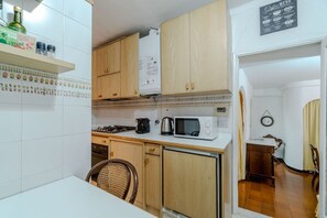 Una cocina compacta con armarios de madera, un microondas, una tetera y un pequeño comedor con una silla.