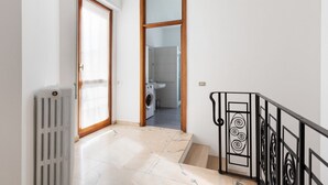 Ein Flur mit Heizkörper, Holztür und Treppe mit schwarzen Geländern, mit Blick in ein Badezimmer.
