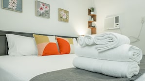Una cama bien hecha con almohadas blancas y naranjas, un cabecero gris y una toalla blanca doblada sobre la cama.