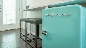 Un refrigerador color turquesa con la palabra 'Frigidaire', un taburete y una ventana con persianas.