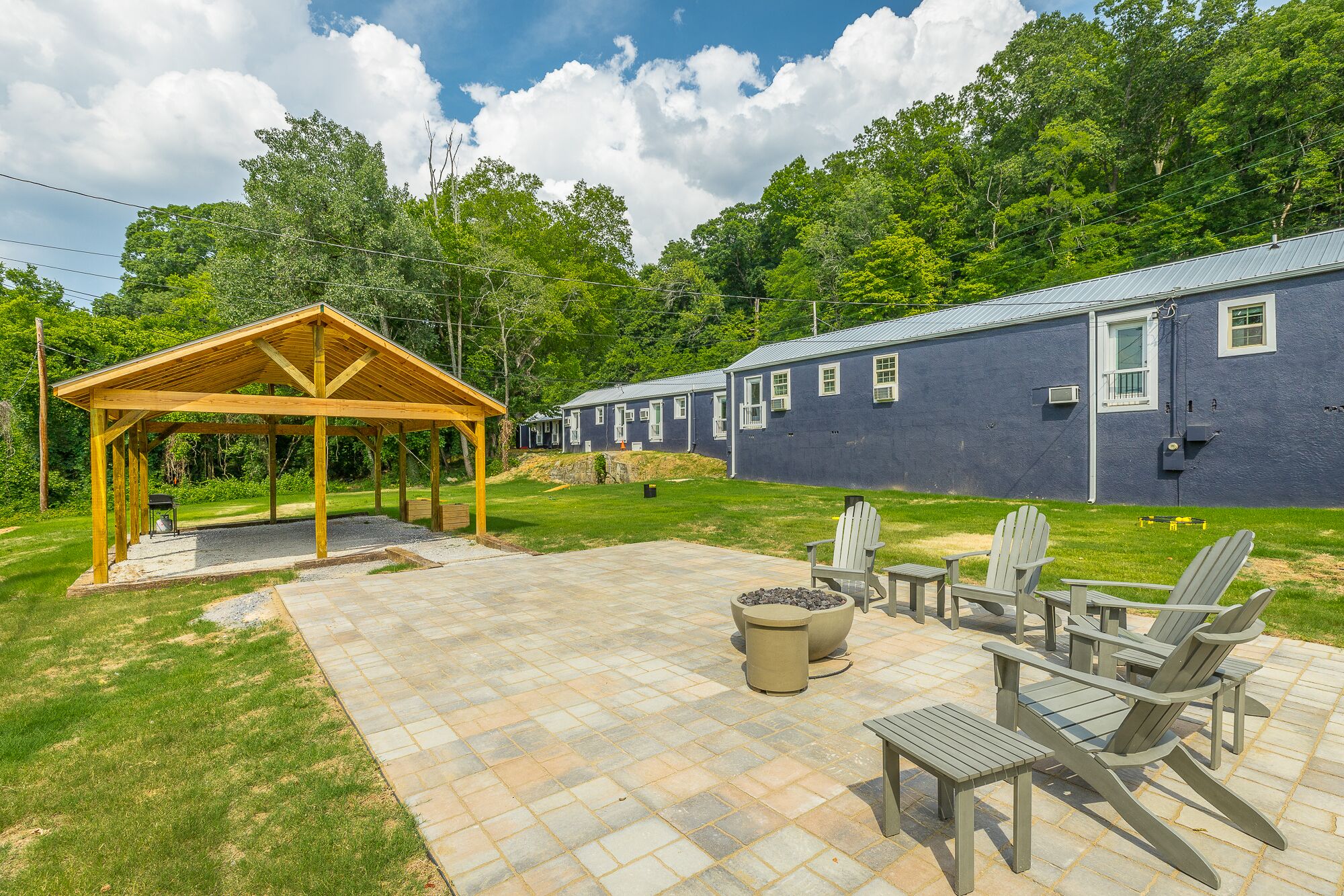 A covered outdoor seating area with Adirondack chairs and a fire pit, adjacent to a row of blue buildings with windows.