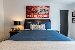 A bedroom with a bed, a nightstand with a lamp, a framed picture on the wall, and a door.