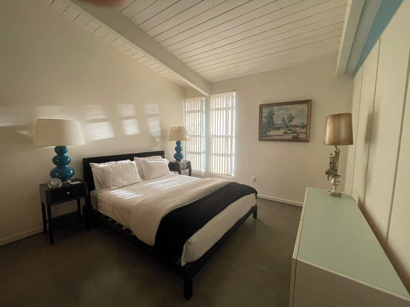 A bedroom with a bed, two bedside tables with lamps, a nightstand, a desk, and a window with blinds.