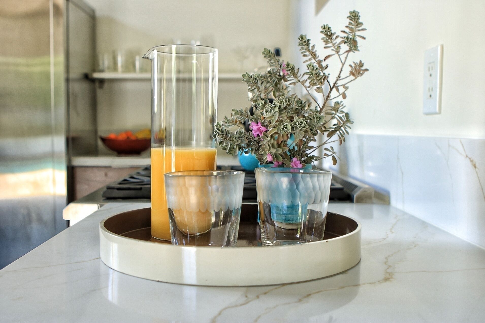 Una encimera de cocina con superficie de mármol, una jarra alta de cristal y dos vasos más pequeños llenos de un líquido amarillo, probablemente zumo o batido.