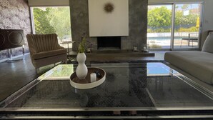A modern living room with a fireplace, two sofas, a coffee table with a vase, and a large window offering a view of a pool and greenery. - MCM Poolside Paradise - Mountain Views & Total Privacy (Palm Springs)