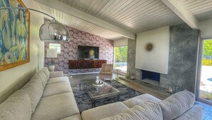 A modern living room with a sectional sofa, a flat-screen TV, a fireplace, and a large window. - MCM Poolside Paradise - Mountain Views & Total Privacy (Palm Springs)