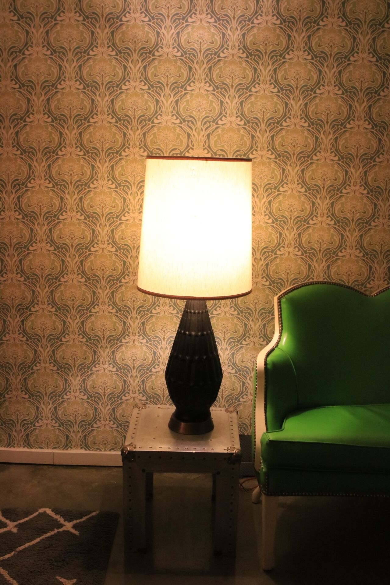 Una lámpara sobre una pequeña mesa de metal junto a una silla verde, con un papel tapiz estampado al fondo.